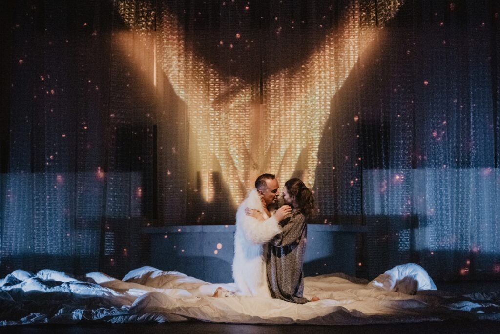 Zdjęcie ze spektaklu Mistrz i Małgorzata. Aktorka i aktor klęczą objęci na pościeli rozłożonej na scenie. Aktorka ma na sobie szlafrok, a aktor futro. Z tyłu na ścianie wyświetlone są dłonie, które się stykają.