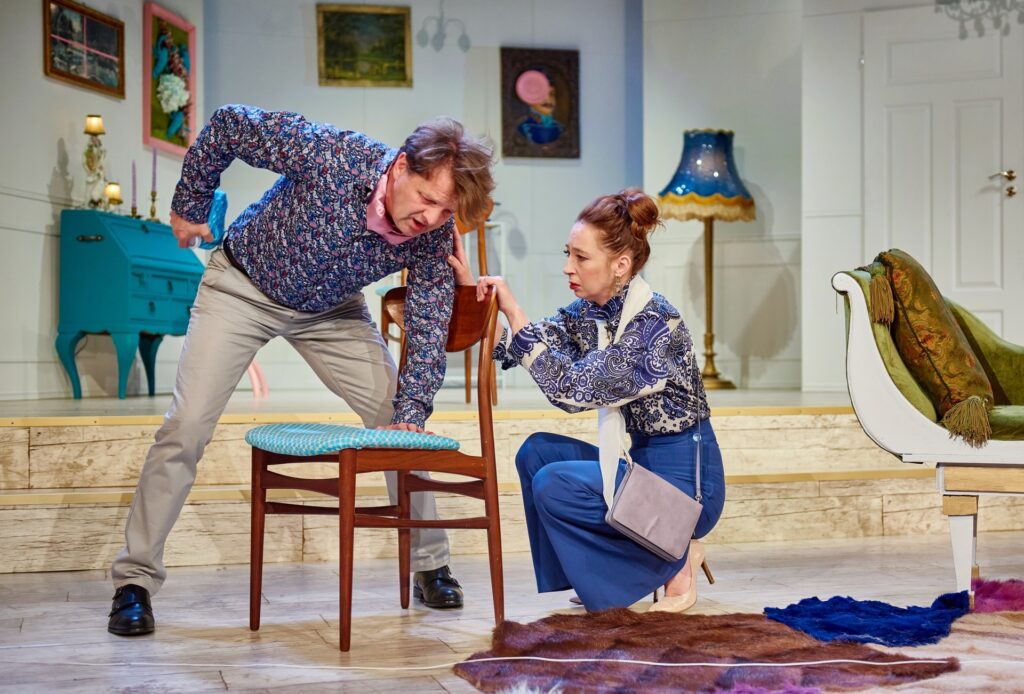 Zdjęcie ze spektaklu Kolacja dla głupca. Aktor pochyla się i opiera jedną dłonią o krzesło. Obok aktorka kuca i trzyma dłońmi oparcie krzesła. W tle jest mieszkanie z awangardowymi, dekoracjami.