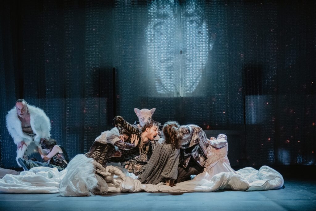 Zdjęcie ze spektaklu Mistrz i Małgorzata. Scena zbiorowa. Aktorzy i aktorki klęczą na scenie, dotykają się, mają różne pozy. Jeden aktor ma głowę kota. Aktor w futrze stoi pochylony z boku. Na ścianie wyświetlona jest twarz mężczyzny z wąsem.