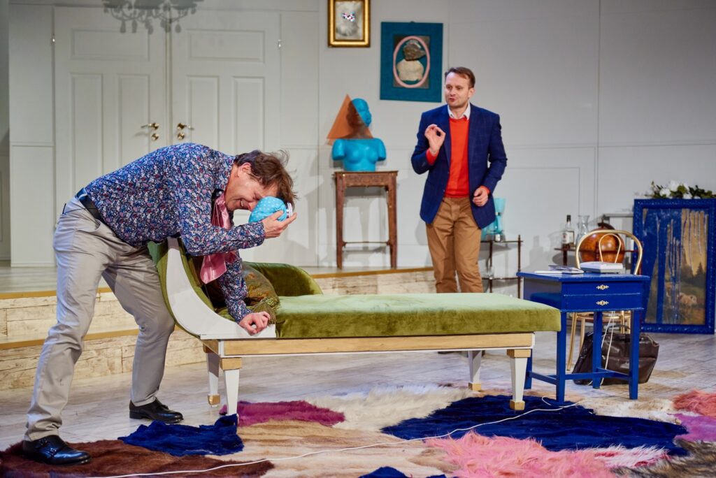 Zdjęcie ze spektaklu Kolacja dla głupca. Aktor opiera się o szezlong, przykłada kompres do czoła. W tle stoi drugi aktor. Scenografia to mieszkanie z awangardowymi dekoracjami. Jest futrzasty dywan, kolorowe meble, rzeźba i obrazy.