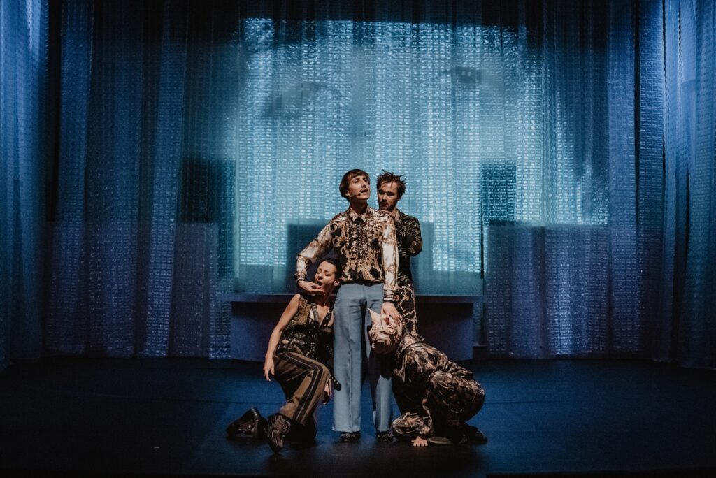 Zdjęcie ze spektaklu Mistrz i Małgorzata. Zdjęcie zabarwione na niebiesko. Na środku stoi młody aktor z wąsem. Obok wije się aktorka. Za nim stoi drugi aktor. Przed nim klęczy aktor z głową kota. Na ścianie wyświetlona jest twarz aktora z wąsem.