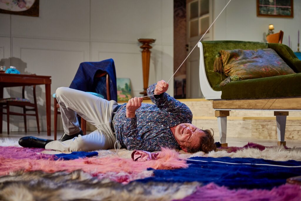 Zdjęcie ze spektaklu Kolacja dla głupca. Aktor leży na futrzastym dywanie, ma wykrzywioną twarz, w ręce trzyma sznurek.