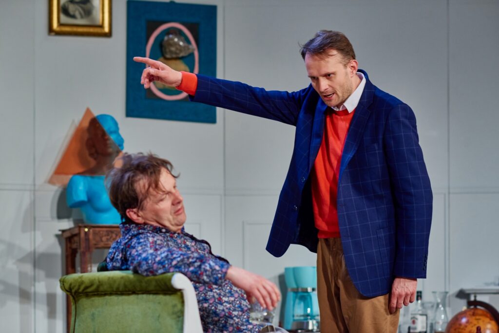 Zdjęcie ze spektaklu Kolacja dla głupca. Aktor siedzi na szezlongu. Nad nim stoi drugi aktor i wskazuje coś palcem. W tle jest rzeźba kobiety bez rąk i obrazy.