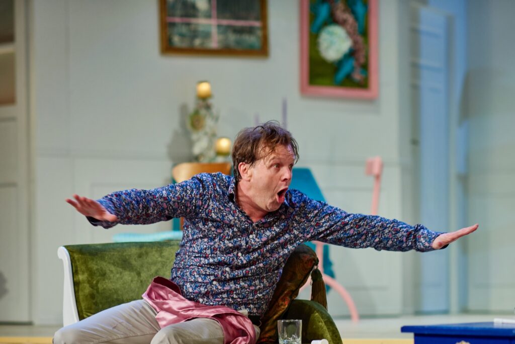 Zdjęcie ze spektaklu Kolacja dla głupca. Aktor siedzi na szezlongu. Patrzy w bok, rozkłada szeroko dłonie i otwiera usta.