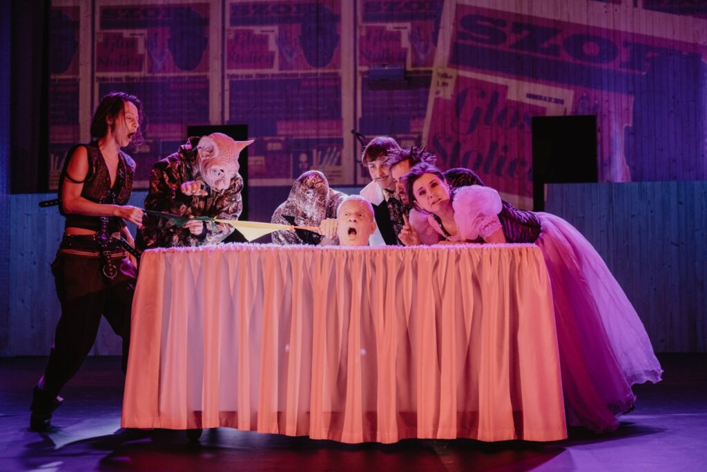 Zdjęcie ze spektaklu Mistrz i Małgorzata. Ze stołu wychodzi głowa aktora. Pozostali aktorzy i aktorki pochylają się nad stołem. Aktor ma głowę kota. Aktorka wyciąga z ucha aktora na stole kolorowe chusteczki. Na ścianie wyświetlane są gazety.