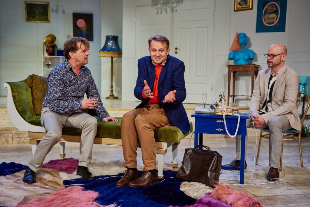 Zdjęcie ze spektaklu Kolacja dla głupca. W mieszkaniu z awangardowymi dekoracjami siedzą dwaj aktorzy na szezlongu. Obok nich jest stolik ze starodawnym telefonem. Z boku na krześle siedzi aktor w garniturze.