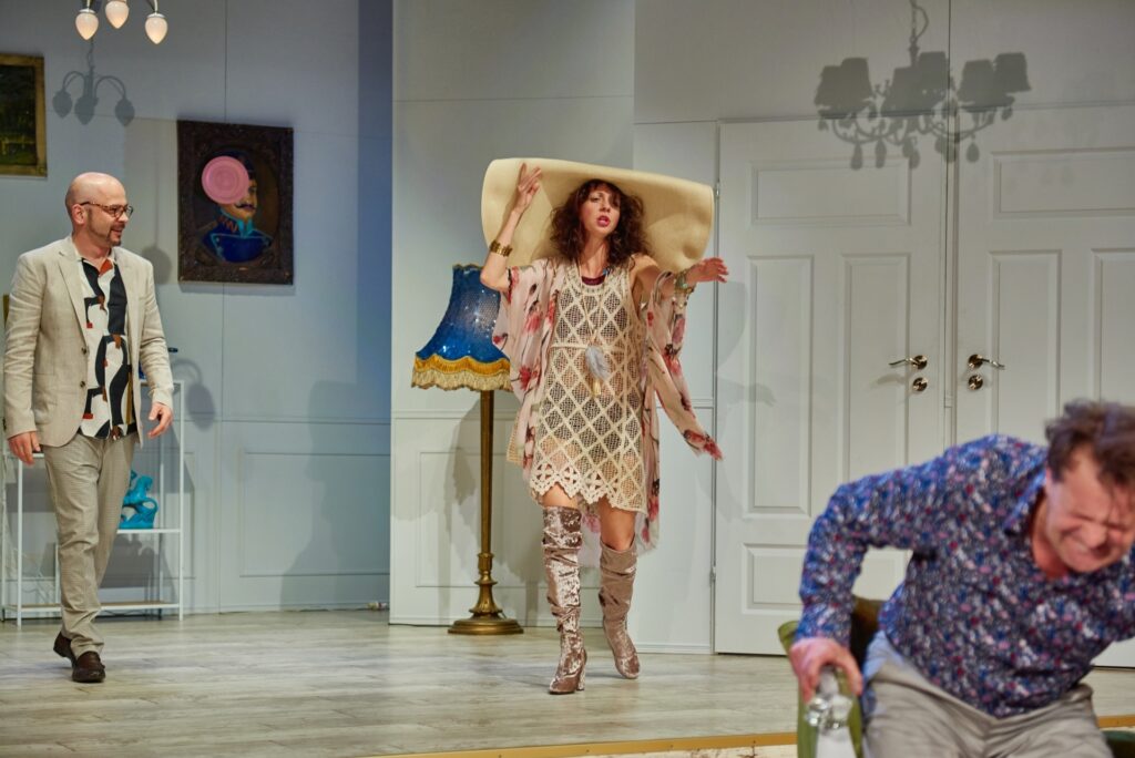 Zdjęcie ze spektaklu Kolacja dla głupca. W rogu aktor próbuje wstać z szezlonga, krzywi się. Z tyłu aktorka wyciąga do niego rękę. Aktorka ma sukienkę siateczkową, narzutę w kwiaty, kozaki i kapelusz. Obok stoi aktor w garniturze.