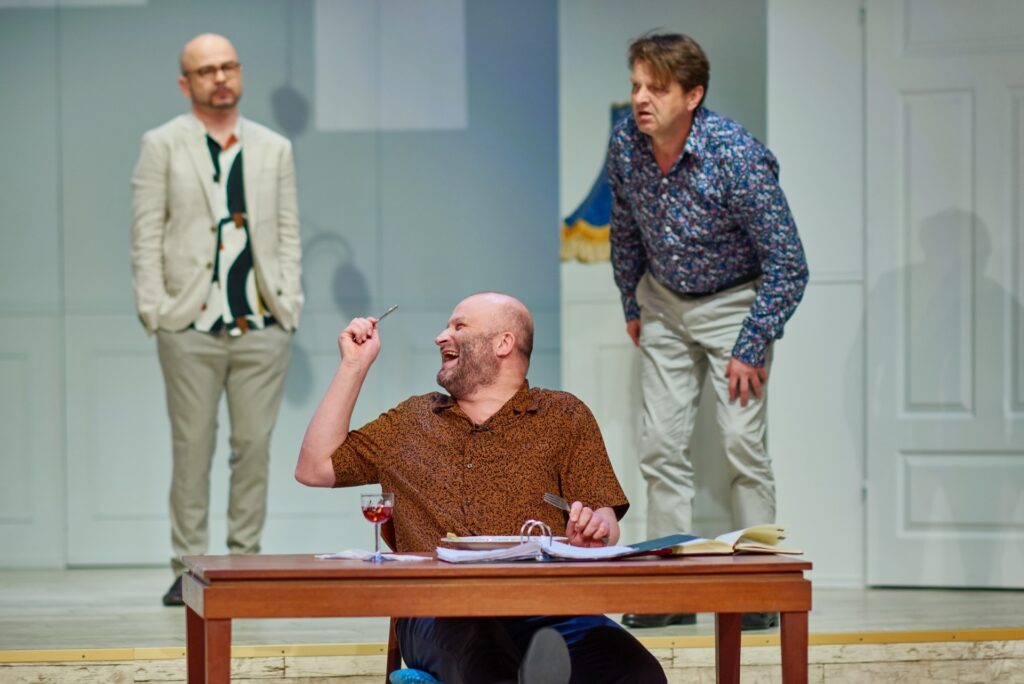 Zdjęcie ze spektaklu Kolacja dla głupca. Łysy aktor siedzi przy stole. Odwraca głowę w bok i się śmieje. W rękach trzyma sztućce. Przed nim na stole jest talerz, kieliszek z winem i segregator z dokumentami. Z tyłu stoi dwóch aktorów.