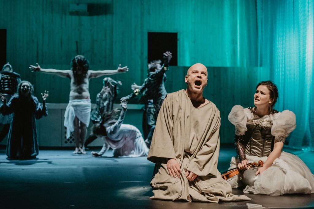 Zdjęcie ze spektaklu Mistrz i Małgorzata. Łysy aktor w stroju pokutnym i aktorka w stroju księżniczki ze skrzypcami klęczą z przodu. W tle aktor w stroju Jezusa, rozkłada ramiona, ma przepaskę na biodrach. Wokół niego są pozostali aktorzy i aktorki.