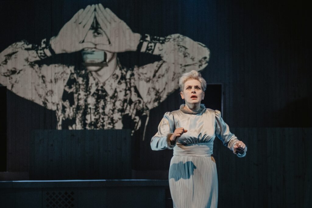 Zdjęcie ze spektaklu Mistrz i Małgorzata. Aktorka w krótkich włosach i długiej sukni. Podnosi ręce, ma zaciśnięte pięści. Jest przestraszona. W tle wyświetlony jest mężczyzna, który ma zakryte usta i przykłada do oka dłonie ułożone w trójkąt