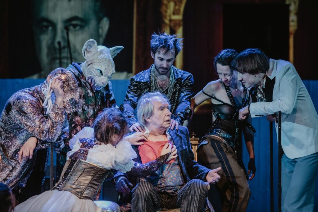 Zdjęcie ze spektaklu Mistrz i Małgorzata. Scena zbiorowa. Starszy aktor siedzi na krześle. Wokół niego stoją pozostali aktorzy i aktorki. Jeden aktor ma głowę kota. Na ścianie wyświetlona jest twarz mężczyzny.