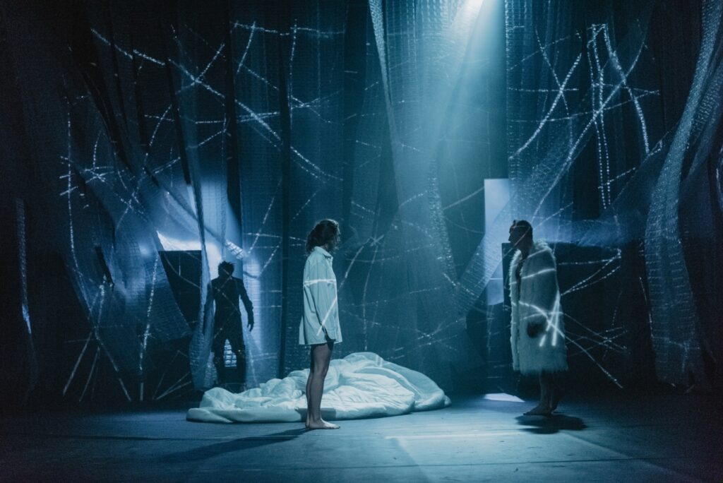 Zdjęcie ze spektaklu Mistrz i Małgorzata. Niebieskie światło, wzory laserowe. Aktorka w białej koszuli i aktor w białym futrze stoją naprzeciwko siebie. Z tyłu stoi inny aktor. W tle jest zwinięte prześcieradło i paskowa kurtyna.