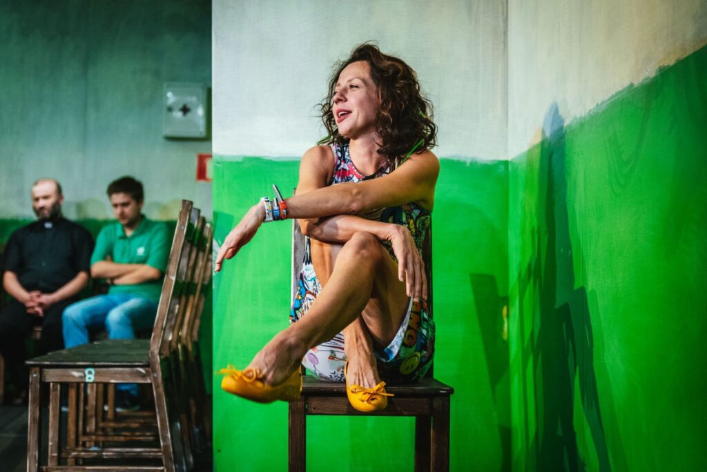 Zdjęcie ze spektaklu Gdy przyjdzie sen. Na pierwszym planie aktorka we wzorzystej sukience siedzi na krześle. Aktorka podciągnęła nogi i ma stopy oparte na krześle. W tle na krzesłach siedzi dwóch aktorów. Jeden ma koszulę z koloratką.