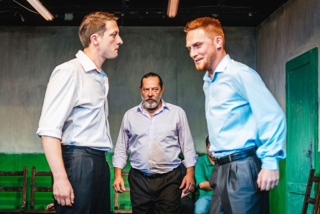 Zdjęcie ze spektaklu Gdy przyjdzie sen. Dwaj młodzi aktorzy stoją naprzeciwko siebie i mierzą się wzrokiem. Między nimi stoi starszy aktor i patrzy na aktora po prawej stronie.