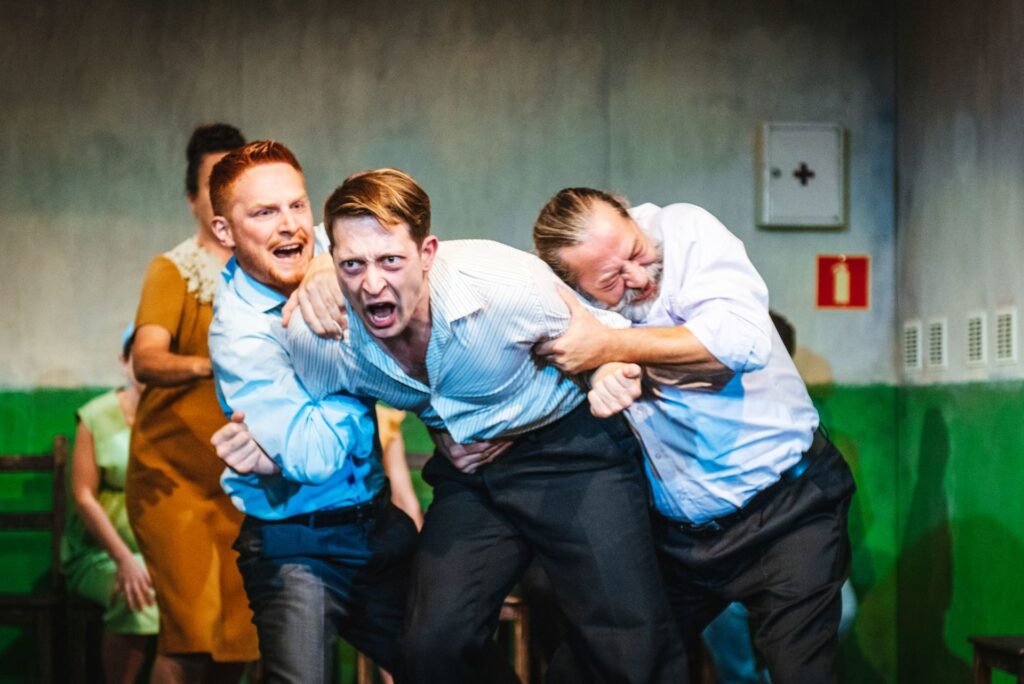 Zdjęcie ze spektaklu Gdy przyjdzie sen. Młody aktor krzyczy i pochyla się do przodu. Drugi młody aktor i starszy aktor trzymają go pod ramiona z dwóch stron. W tle jedna aktorka podchodzi do mężczyzn, a druga siedzi na krześle.