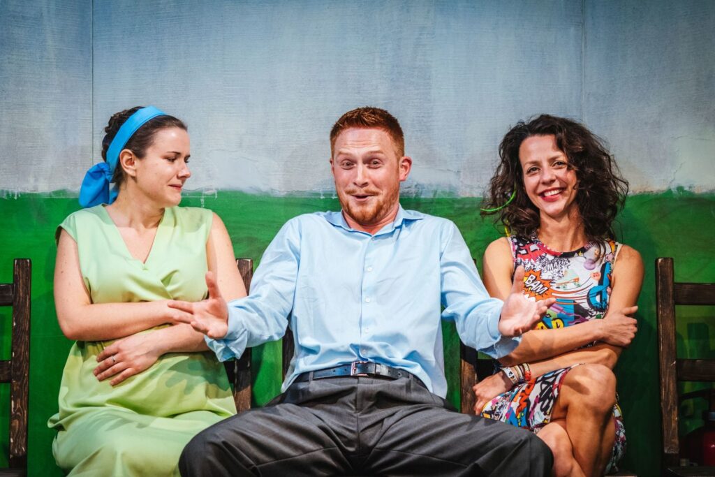 Zdjęcie ze spektaklu Gdy przyjdzie sen. Na krzesłach pod ścianą siedzi aktor i dwie aktorki. Aktor rozkłada ręce na boki. Jedna aktorka patrzy na aktora i trzyma się za brzuch ciążowy. Druga aktorka patrzy na wprost i się uśmiecha.