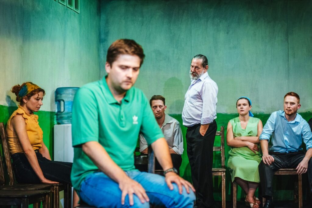 Zdjęcie ze spektaklu Gdy przyjdzie sen. Na krześle siedzi aktor, przymyka oczy i trzyma dłonie na kolanach. Pod ścianą siedzą pozostali aktorzy i aktorki. Starszy aktor stoi i patrzy na aktora na pierwszym planie. W kącie stoi dozownik wody.