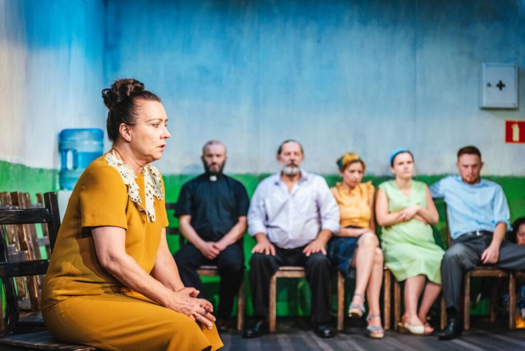 Zdjęcie ze spektaklu Gdy przyjdzie sen. Na pierwszym planie bokiem siedzi aktorka w średnim wieku. Ma sukienkę i włosy upięte w koka. Pod ścianą siedzą pozostali aktorzy i aktorki. Jeden aktor ma koszulę z koloratką. W kącie stoi dozownik wody.