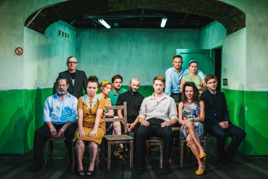 Zdjęcie ze spektaklu Gdy przyjdzie sen. Zdjęcie grupowe. Wszyscy aktorzy i aktorki siedzą na krzesłach lub stoją.