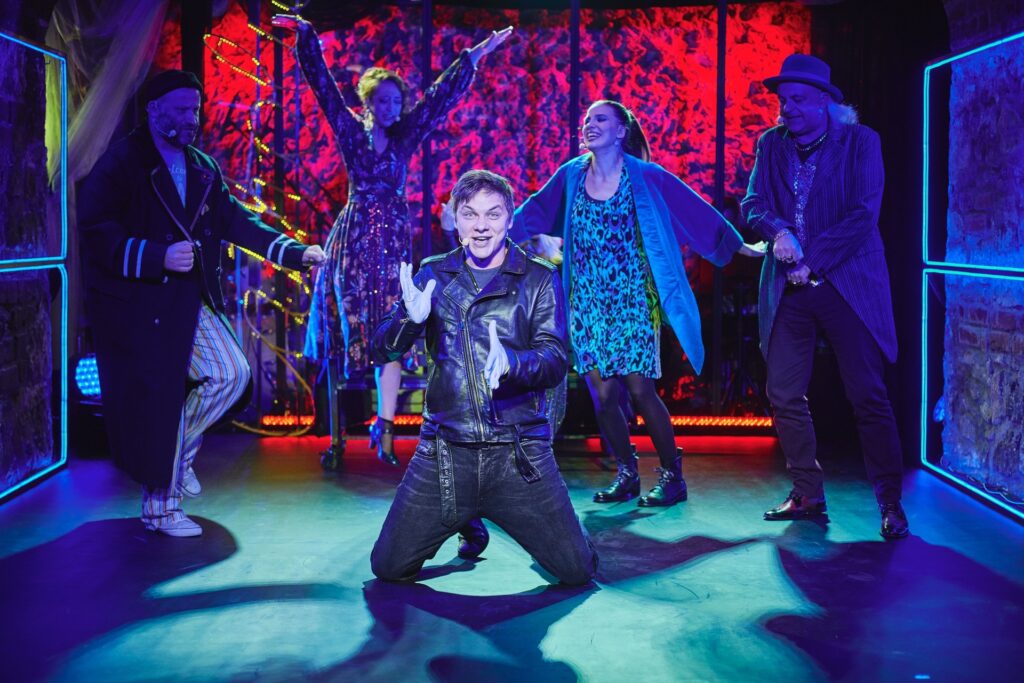 Zdjęcie ze spektaklu Krakowska lekkość bytu. Aktor w skórzanej kurtce i białych rękawiczkach klęczy na środku. Za nim pozostali aktorzy i aktorki tańczą i skaczą. Scena jest oświetlona w dwóch kontrastujących kolorach, czerwonym i niebieskim.