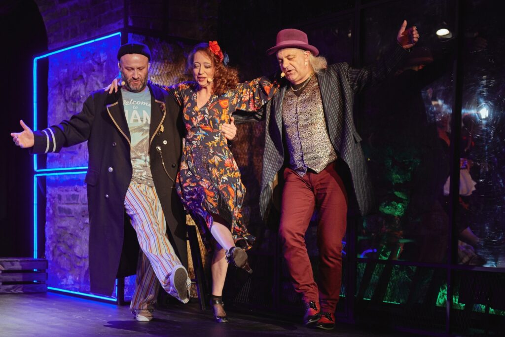 Zdjęcie ze spektaklu Krakowska lekkość bytu. Dwóch aktorów i aktorka trzymają się za ramiona i podnoszą nogi jakby tańczyli kankana. Aktorka jest w środku. Wszyscy mają kolorowe ekstrawaganckie stroje.