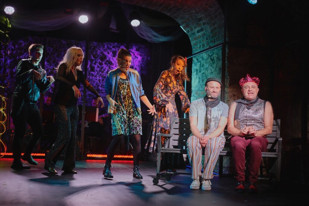 Zdjęcie ze spektaklu Krakowska lekkość bytu. Z boku na ławce siedzi dwóch aktorów, mają szaliki i czapki. Jeden z nich ma błyszczącą czapkę. Od tyłu skradają się do nich trzy aktorki i aktor.