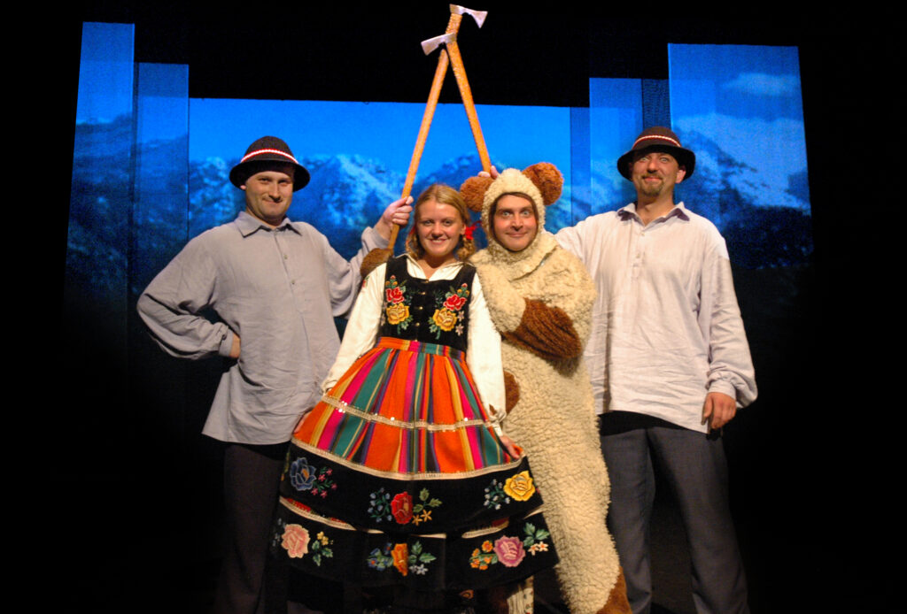 Zdjęcie ze spektaklu Pyza na polskich dróżkach. Na środku stoi aktorka z warkoczami w łowickim stroju ludowym. Aktor w stroju misia trzyma aktorkę za ramiona. Z boku stoją aktorzy w koszulach, czapkach góralskich i unoszą ciupagi.