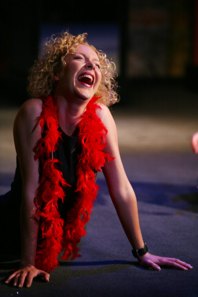 Zdjęcie ze spektaklu Wszystko o kobietach. Aktorka z kręconymi włosami siedzi na scenie. Podpiera się rękami. Na szyi ma szal z piór.