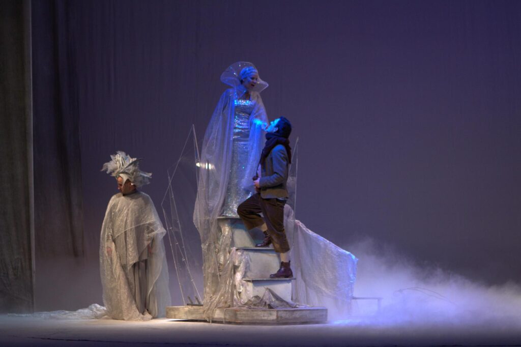 Zdjęcie ze spektaklu Królowa Śniegu. Na piedestale stoi kobieta ubrana na biało, na schodkach podchodzi do niej chłopiej w zimowym ubraniu. Z boku stoi postać w białych szatach zakrywających sylwetkę. Scena jest pusta z zadymieniem.