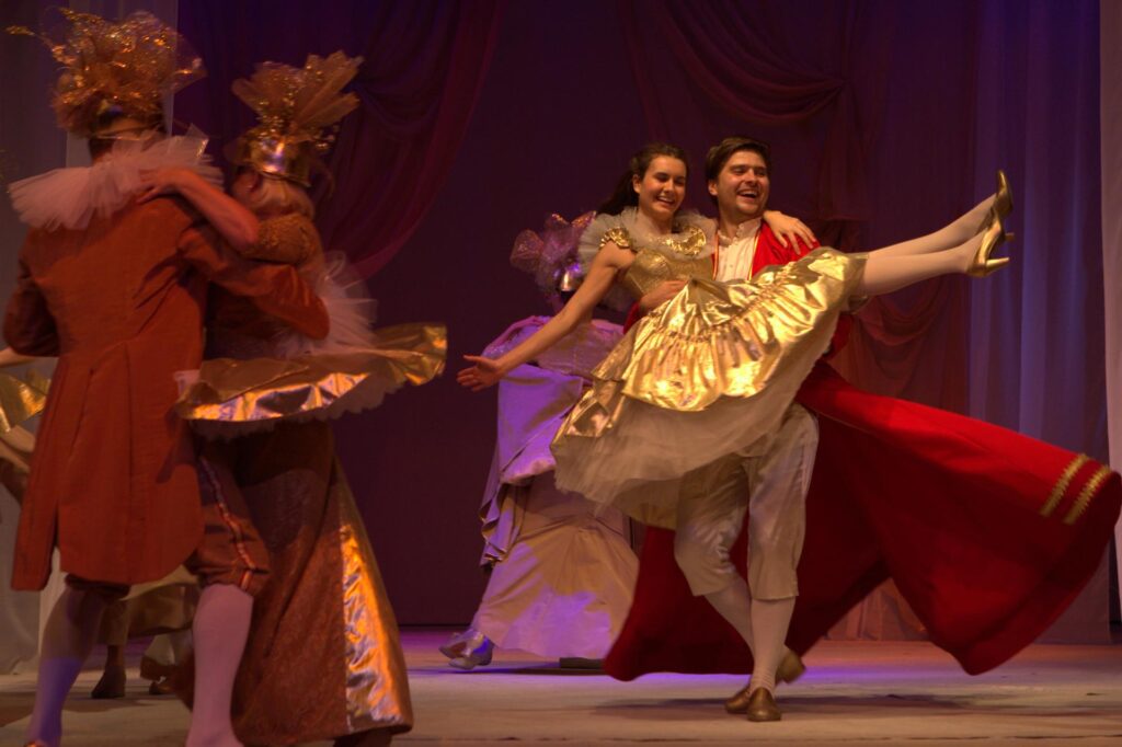 Zdjęcie ze spektaklu Królowa Śniegu. Scena zbiorowa, tańczące postacie w maskach. Postać księcia ma na rękach dziewczynę w balowej sukni.