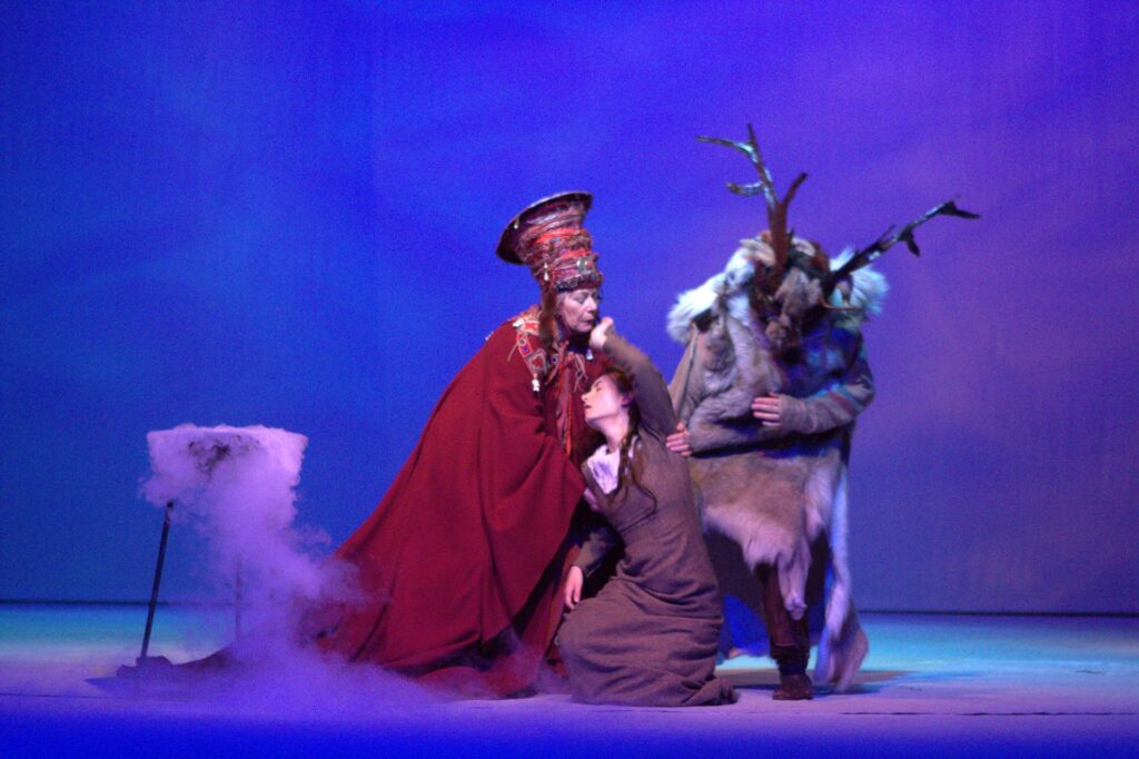 Zdjęcie ze spektaklu Królowa Śniegu. Na pierwszym planie kobieta w stroju etnicznym lapońskim, podtrzymuje omdlewającą dziewczynę w szarej sukience. Obok postać w futrze przebrana za renifera.