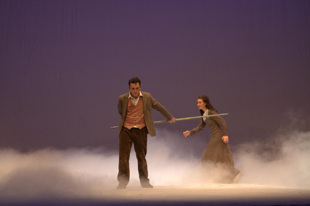 Zdjęcie ze spektaklu Królowa Śniegu. Scena jest bez dekoracji z zadymieniem. Na pierwszym planie stoi chłopiec wymachujący długim kijem, za nim w biegu dziewczynka w szarej sukience.