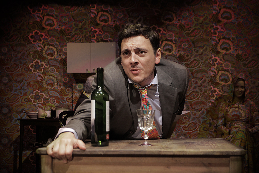 Zdjęcie ze spektaklu Sarenki. Zbliżenie. Aktor w garniturze siedzi i trzyma brzeg stołu. Na stole jest kieliszek i butelka wina. Przy ścianie siedzi aktorka. Sukienka aktorki i krawat aktora mają taki sam wzór jak tapeta na ścianie