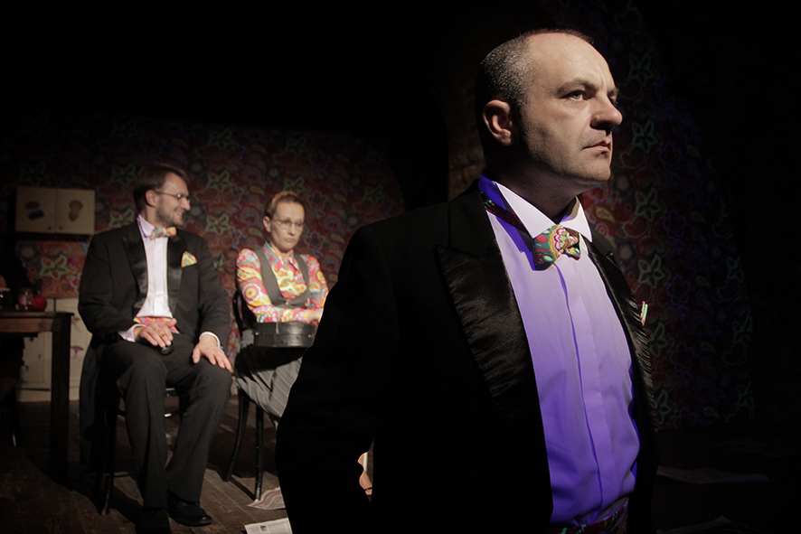 Zdjęcie ze spektaklu Sarenki. Zbliżenie. Aktor w smokingu patrzy przed siebie. Za nim siedzi aktor w smokingu i okularach oraz aktorka w spódnicy i kamizelce. Akcesoria aktorów i aktorki mają taki sam wzór jak tapeta na ścianie.