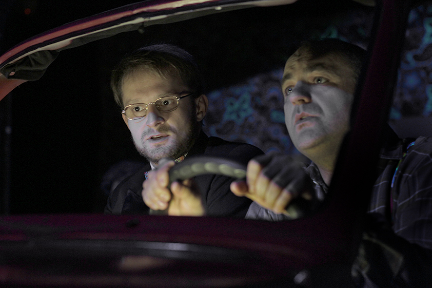 Zdjęcie ze spektaklu Sarenki. Zbliżenie. Aktor w koszuli w kratę i aktor w smokingu i okularach siedzą w samochodzie. Aktor w koszuli w kratę trzyma kierownicę.