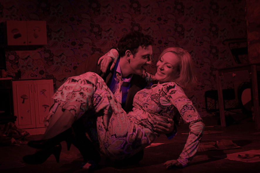 Zdjęcie ze spektaklu Sarenki. Zdjęcie zabarwione na czerwono. Aktor w garniturze klęczy i podnosi aktorkę. Sukienka aktorki i krawat aktora mają taki sam wzór jak tapeta na ścianie. Na podłodze są gazety.