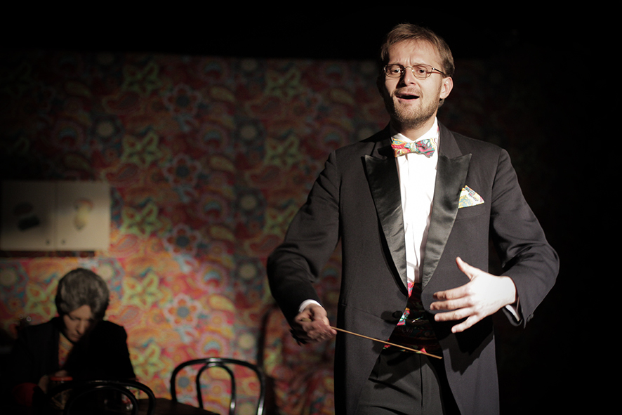 Zdjęcie ze spektaklu Sarenki. Aktor w smokingu i okularach otwiera usta, w dłoni trzyma batutę. Z tyłu przy stole siedzi aktorka. Pas, mucha i poszetka aktora mają taki sam wzór jak tapeta na ścianie.