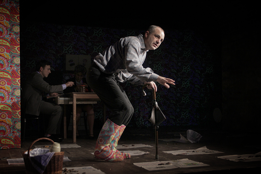 Zdjęcie ze spektaklu Sarenki. Aktor w koszuli w kratę i we wzorzystych materiałowych kaloszach opiera się na parasolce. Na scenie leżą gazety. Z tyłu przy stole siedzi drugi aktor. Na ścianie jest tapeta, tapeta ma taki sam wzór jak kalosze.