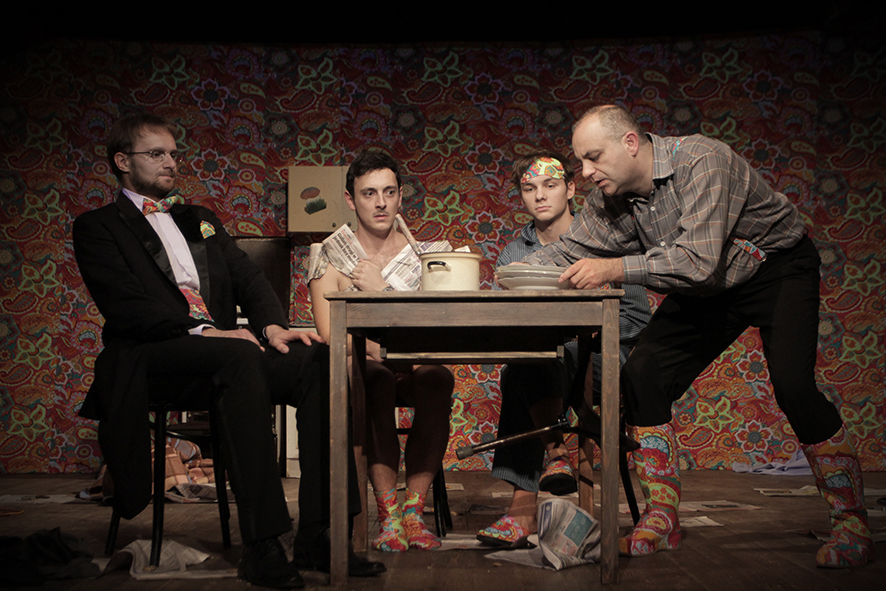 Zdjęcie ze spektaklu Sarenki. Trzech aktorów siedzi przy stole. Jeden jest w smokingu, drugi nie ma koszulki i zasłania się gazetami, trzeci ma opaskę. Inny aktor stawia na stole talerze. Akcesoria aktorów mają taki sam wzór jak tapeta na ścianie