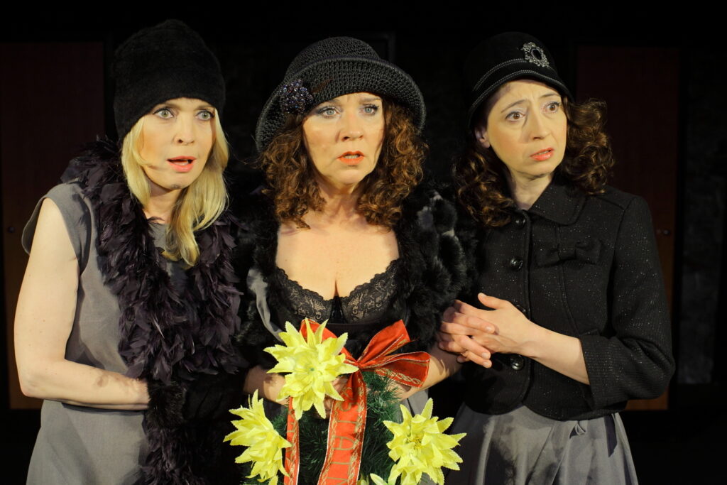 Zdjęcie ze spektaklu Wszystko o kobietach. Trzy aktorki stoją obok siebie i trzymają się pod rękę. Mają zdziwione miny. Są ubrane na czarno, na głowach mają kapelusze. Aktorka w środku trzyma wieniec pogrzebowy.