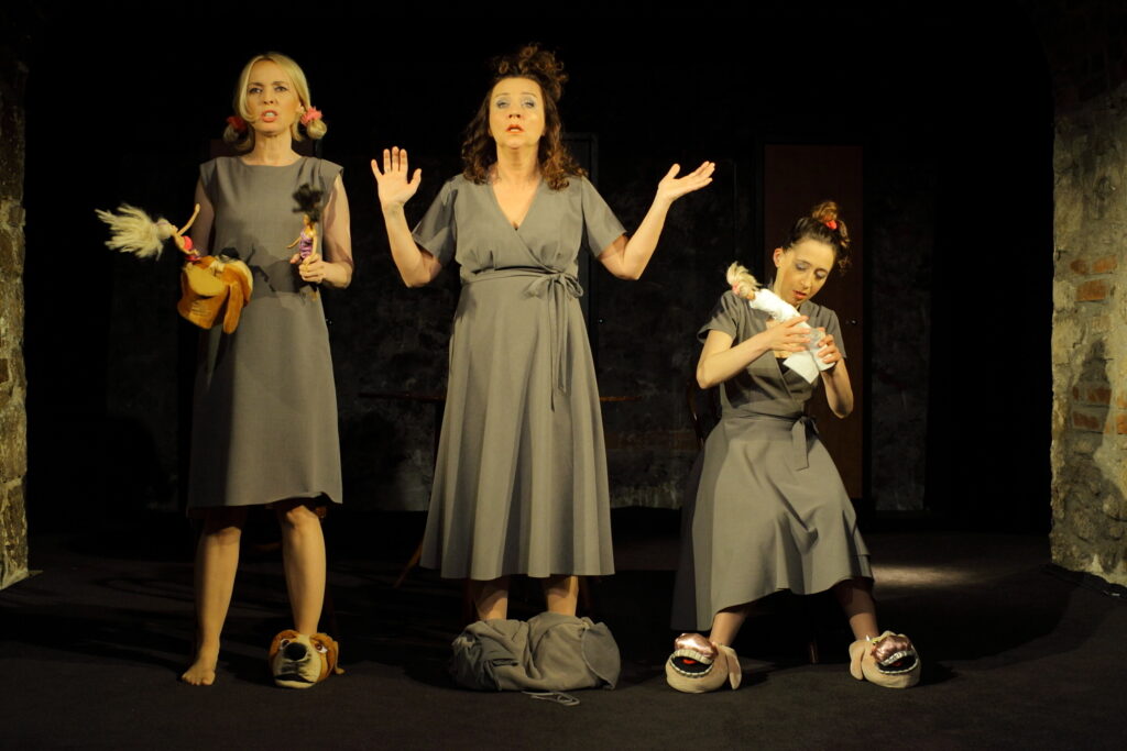 Zdjęcie ze spektaklu Wszystko o kobietach. Trzy aktorki w sukienkach. Mają włosy związane w kucyki, na stopach mają kapcie miśki. Aktorka na środku rozkłada ręce, stopy ma przykryte kocem. Aktorki po bokach trzymają lalki Barbie.