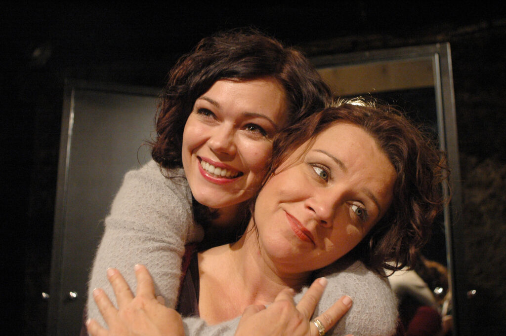 Zdjęcie ze spektaklu Wszystko o kobietach. Zbliżenie. Jedna aktorka obejmuje drugą za szyję od tyłu.