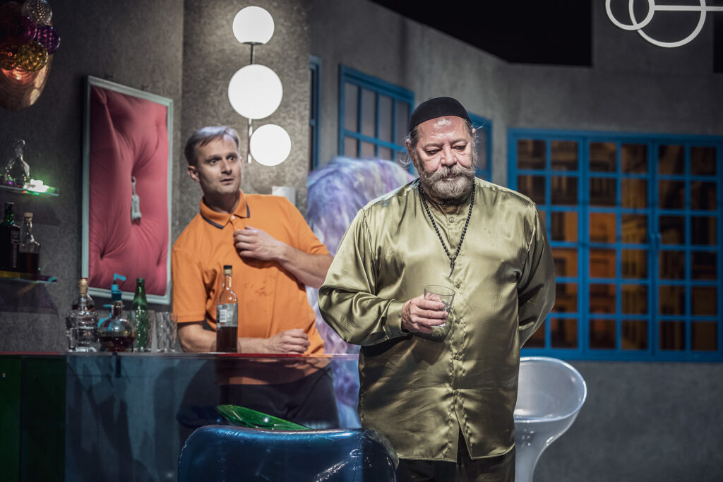 Zdjęcie ze spektaklu Pignon i urząd skarbowy. Starszy aktor z zarostem przymyka oczy, w dłoni ma szklankę. Za nim aktor opiera się o szklany bar, jedną rękę przykłada do klatki piersiowej. W tle są okna i awangardowe dekoracje.