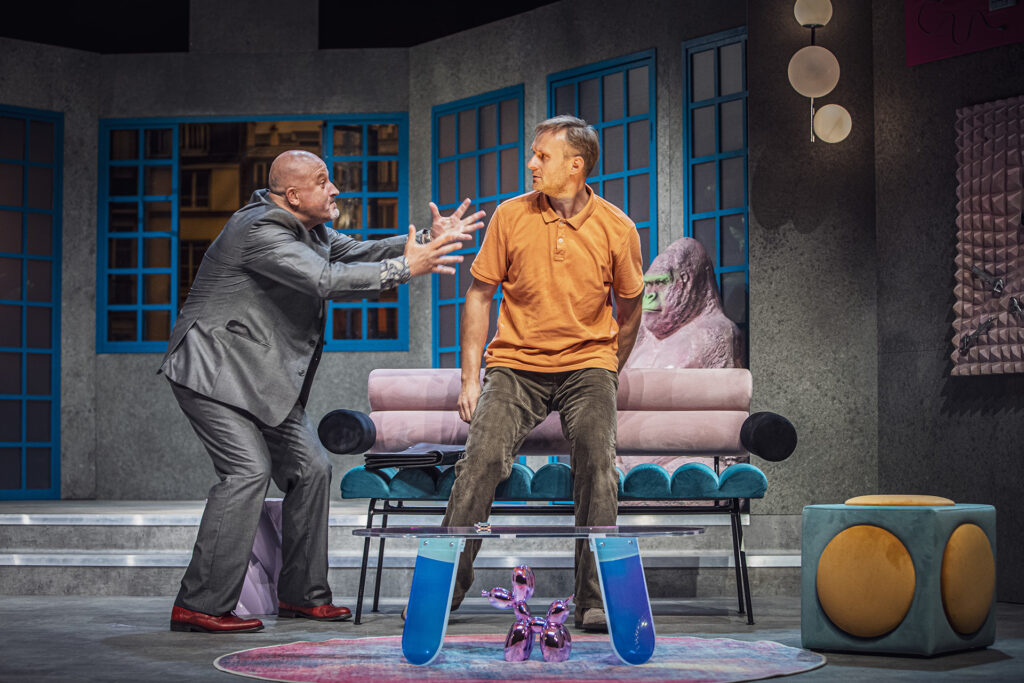 Zdjęcie ze spektaklu Pignon i urząd skarbowy. Aktor wstaje z kanapy. Obok niego stoi łysy aktor i wymachuje rękami. Są w mieszkaniu z awangardowymi dekoracjami. W tle jest rzeźba goryla i okna.