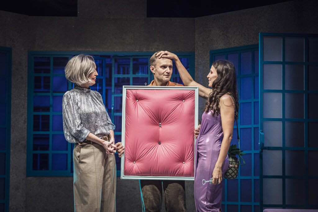 Zdjęcie ze spektaklu Pignon i urząd skarbowy. Aktor trzyma przed sobą ramę do obrazu wypełnioną różowym materiałem. Po jego dwóch stronach stoją dwie aktorki. Jedna aktorka kładzie aktorowi dłoń na głowie. W tle są okna.