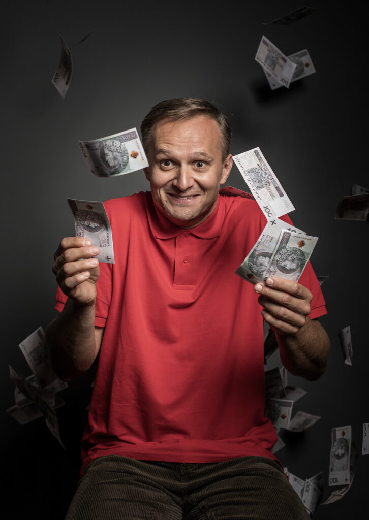 Zdjęcie ze spektaklu Pignon i urząd skarbowy. Portret. Aktor w czerwonej polówce na czarnym tle. Uśmiecha się, w dłoniach trzyma banknoty. Nad nim latają liczne banknoty.