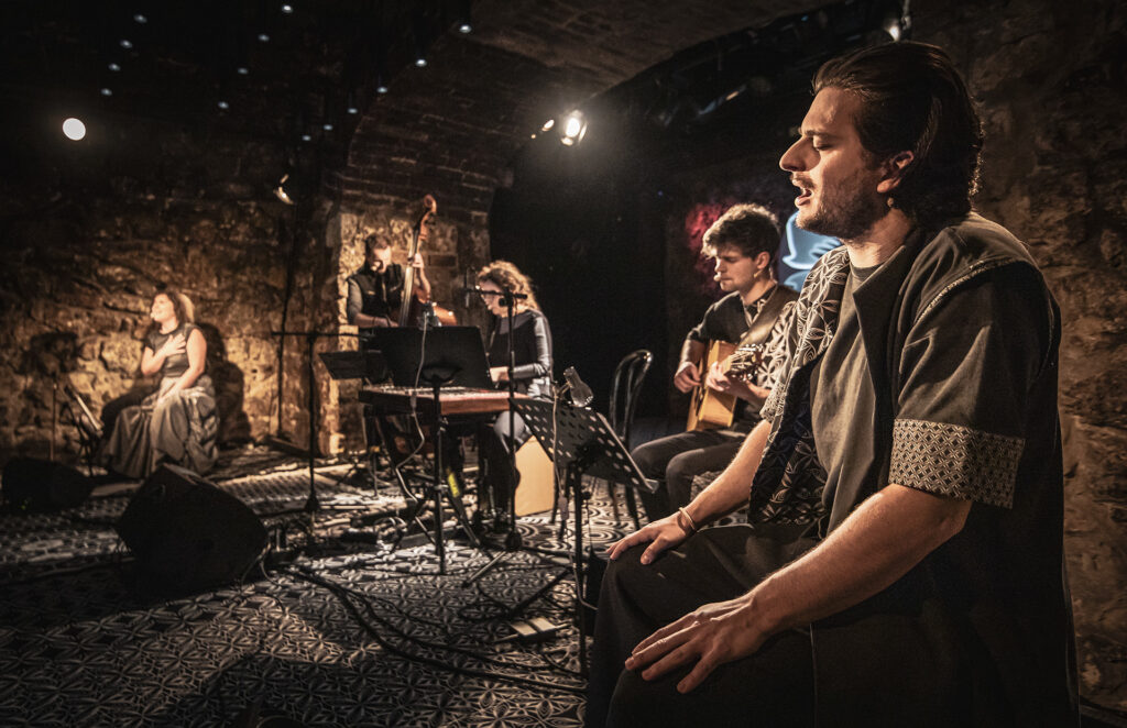 Zdjęcie ze spektaklu Śpiewaj mi, bo chcę. Mężczyzna z włosami do ramion ma zamknięte oczy i śpiewa. W tle siedzi mężczyzna z gitarą i kobieta przy cymbałach. Za nimi stoi mężczyzna z kontrabasem. Obok siedzi kobieta, trzyma rękę na piersi i śpiewa.