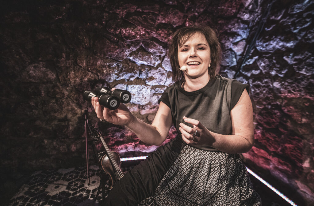 Zdjęcie ze spektaklu Śpiewaj mi, bo chcę. Młoda kobieta w stroju romskim. Uśmiecha się, śpiewa, w ręce trzyma grzechotkę. Za nią na podłodze leżą skrzypce. Wokół są ściany z kamienia.