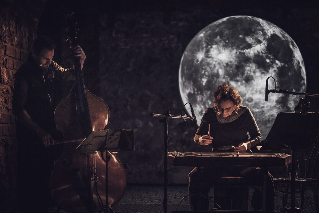 Zdjęcie ze spektaklu Śpiewaj mi, bo chcę. Na scenie kobieta gra na cymbałach. Obok po lewej stronie mężczyzna gra na kontrabasie. Za nimi na ścianie jest wyświetlony księżyc.