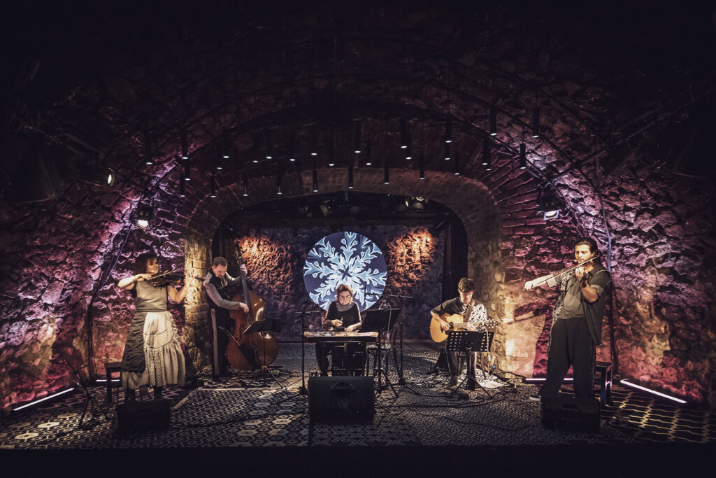 Zdjęcie ze spektaklu Śpiewaj mi, bo chcę. Na scenie są dwie kobiety i trzech mężczyzn. Z lewej skrzypaczka i kontrabasista, na środku kobieta przy cymbałach, z prawej gitarzysta i skrzypek. W tle wyświetlone etniczne wzory romskie.